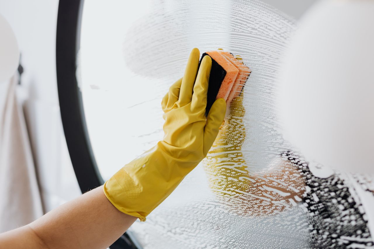 about-us-01 Personne anonyme portant des gants en caoutchouc jaunes et utilisant une éponge simple pour nettoyer un miroir dans une salle de bain